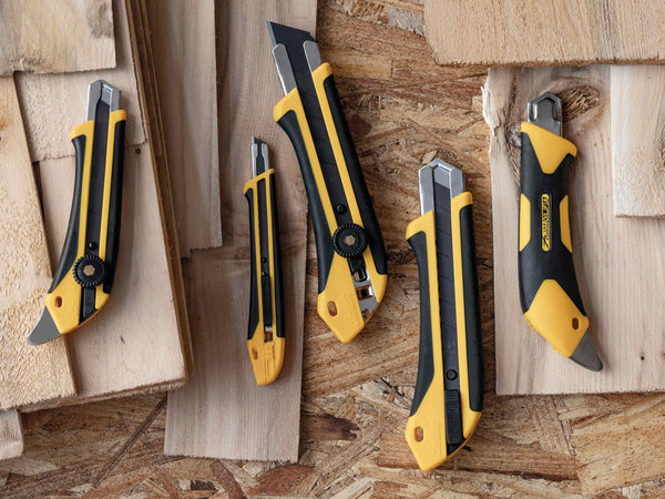 OLFA X-Design Family of Knives on a plank/wooden background. Includes OLFA 18mm L-5, 9mm, XA-1, 25mm XH-1, 25mm XH-AL, and 18mm LA-X.
